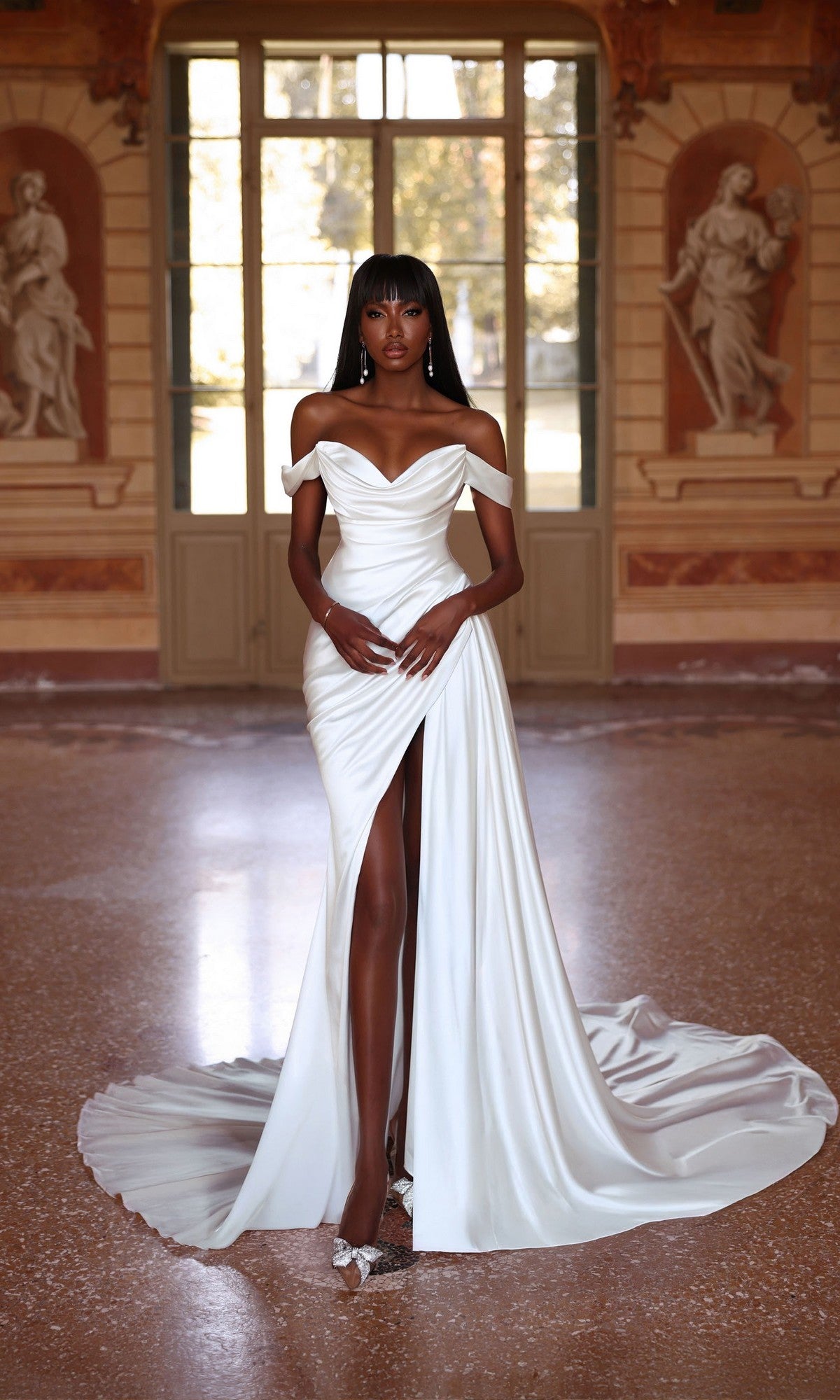 A woman poses in front of tall windows and classical wall sculptures, wearing the Alyce 7106 long ivory dress with a cowl neckline and thigh-high slit.